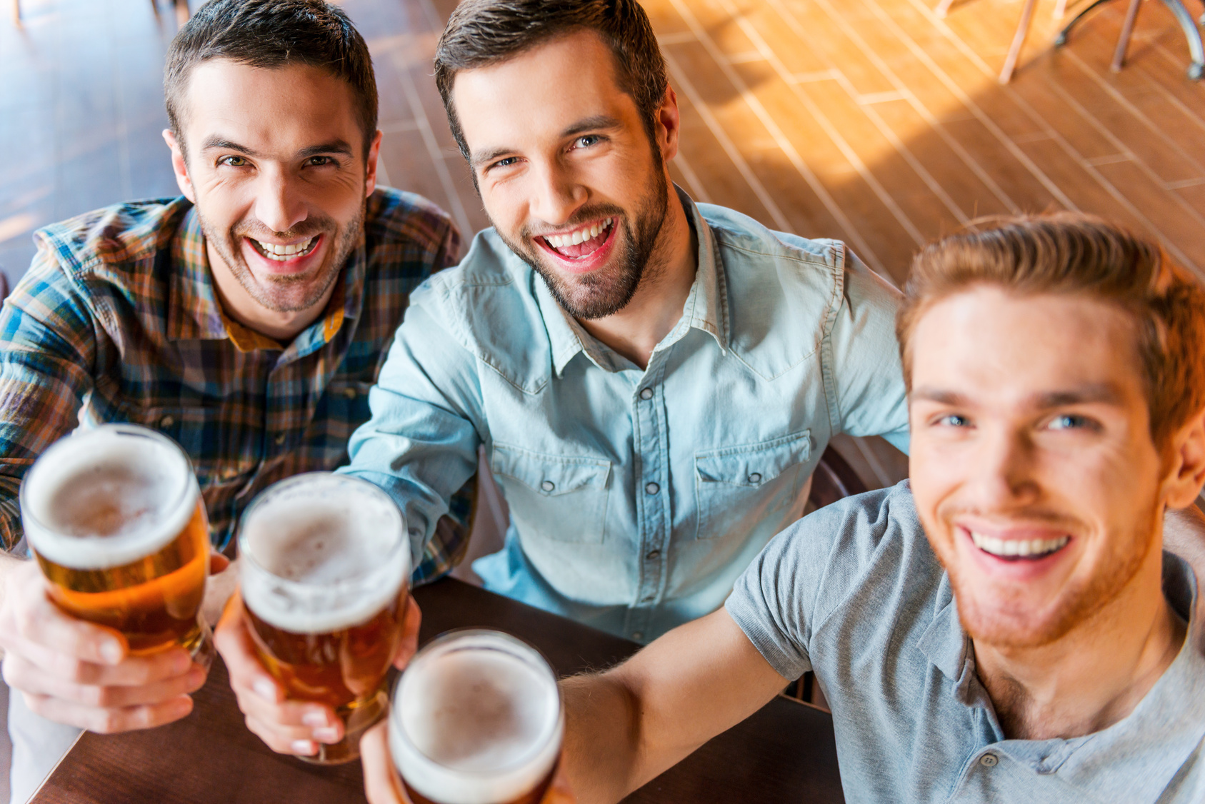 Cheers! Top view of three happy young men in casual wear toasting with beer while sitting in bar together