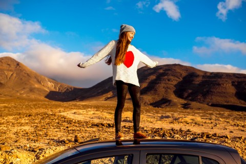 Young carefree woman dressed casual enjoying rocky desert landscape standing on the car roof with raised hands on Fuerteventura island in Spain