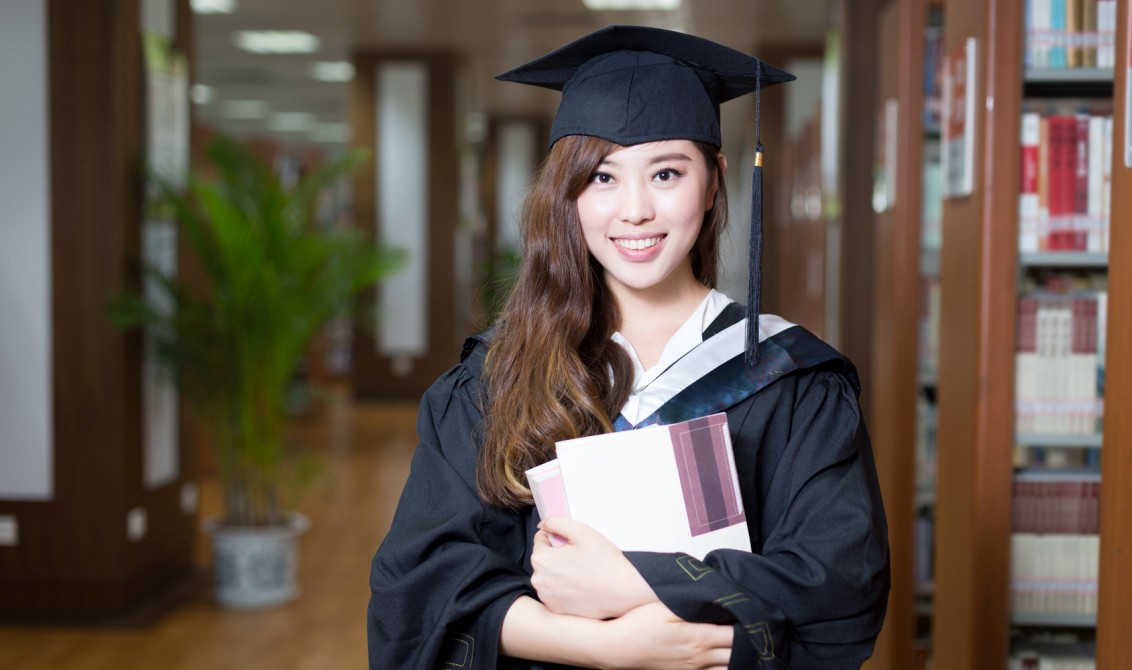 asian female student holding book and wearing academic dress in library