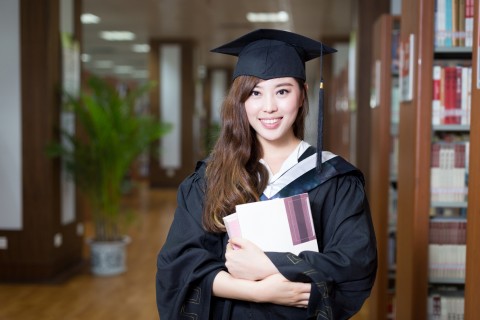 asian female student holding book and wearing academic dress in library